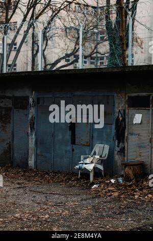 Gebrochener weißgrauer Stuhl vor dem blauen alten Rostiges Garagentor mit Stacheldrahtzaun oben Stockfoto