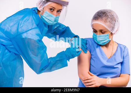 Eine Krankenschwester in Schutzausrüstung, in einer medizinischen Maske, macht eine junge Ärztin einen Impfstoff gegen covid 19 und schaut auf die Kamera. Weißer Hintergrund. Stockfoto