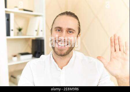 Das Porträt eines attraktiven jungen bärtigen Hipster-Mannes lächelt, winkt und schaut auf die Kamera, Social-Media-Blogger grüßt seine Anhänger Stockfoto