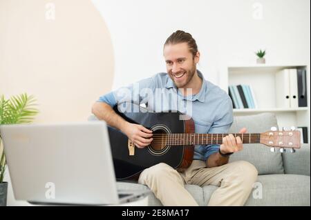 Attraktive fröhlich bärtigen Hipster Mann lächeln und Gitarre spielen, sitzen im Wohnzimmer auf einer bequemen Couch, geben Online-Musikunterricht oder Stockfoto