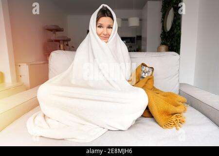 Frau, die sich kalt fühlt. Frau und Katze wärmen sich unter einer Decke auf dem Sofa zu Hause. Frost vor Kälte Stockfoto