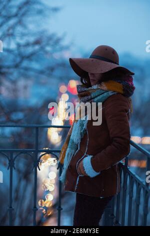 Nachdenklich elegante Frau mittleren Alters in braunem Hut und Schal mit Fäustlingen in Schaffell Mantel in der Nacht draußen im Stadtpark im Winter. Stockfoto