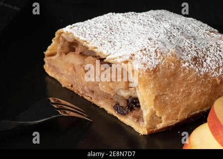 Ein Apfelstrudel auf schwarzem Teller auf schwarzem Hintergrund mit ein paar Apfelscheiben auf der Seite, mit Puderzucker überzogen, geschnitten, zeigt die Füllung Stockfoto