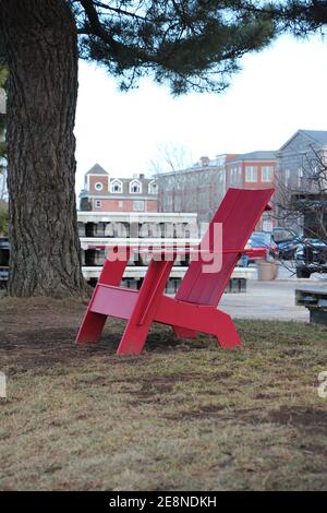 Leuchtend roter Adirondack-Stuhl in einem Park neben einer Kiefer mit Stadtlandschaft im Hintergrund Stockfoto