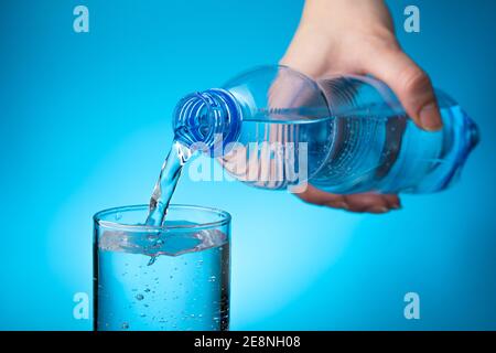 Eine Frauenhand hält eine Plastikflasche und gießt Wasser in ein Glas auf hellblauem Hintergrund. Stockfoto