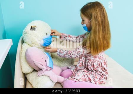 Kleine Mädchen spielen zu Hause während Lockdown Isolation, trägt blaue chirurgische Gesichtsmaske auf sich selbst und Stofftiere Stockfoto