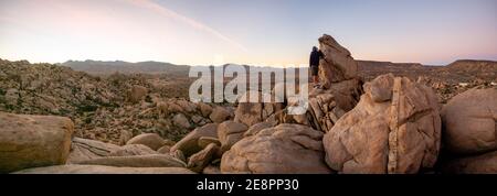Panorama des Mannes Wandern zwischen Feld von Felsbrocken in der Dämmerung mit Sonnenuntergang Himmel Yucca Valley, Kalifornien in der Nähe Joshua Tree National Park an einem sonnigen Januartag Stockfoto