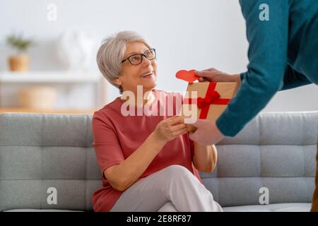 Ein älteres Paar am valentinstag. Fröhliche nette ältere Frau lächelt, während sie ein Geschenk von ihrem Mann erhält. Stockfoto