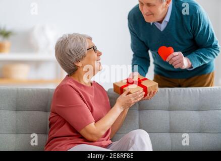 Ein älteres Paar am valentinstag. Fröhliche nette ältere Frau lächelt, während sie ein Geschenk von ihrem Mann erhält. Stockfoto