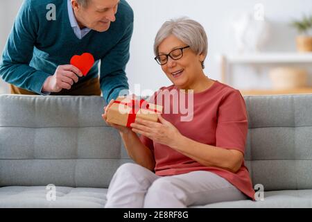 Ein älteres Paar am valentinstag. Fröhliche nette ältere Frau lächelt, während sie ein Geschenk von ihrem Mann erhält. Stockfoto