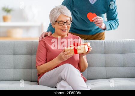 Ein älteres Paar am valentinstag. Fröhliche nette ältere Frau lächelt, während sie ein Geschenk von ihrem Mann erhält. Stockfoto