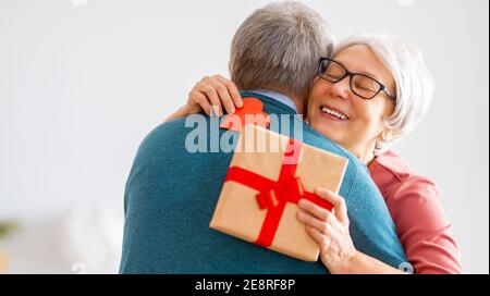 Ein älteres Paar am valentinstag. Fröhliche nette ältere Frau lächelt, während sie ein Geschenk von ihrem Mann erhält. Stockfoto