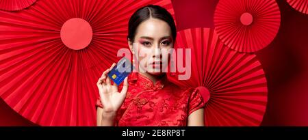 Portrait der schönen asiatischen Frau trägt traditionelle Cheongsam Qipao Kleid Zeigt Kreditkarte in der Hand auf rotem orientalischem Banner Hintergrund Für Chinesisch Stockfoto