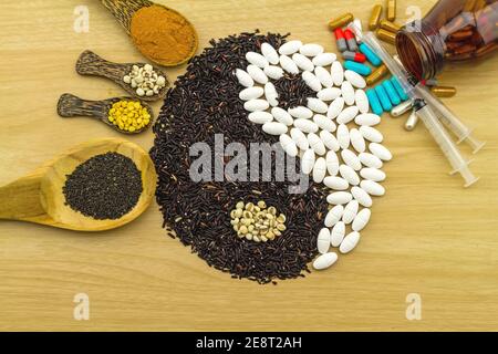 Schwarzer Reis und weiße Pille bilden ein Yin Yang Symbol und Kurkuma-Pulver, Hirse, Sojabohne, Basilikum Samen in Holzlöffel, Spritze, Flasche auf braun w Stockfoto
