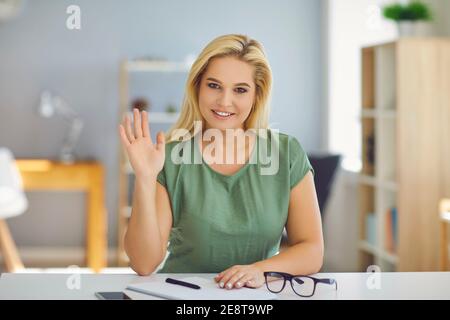 Schöne blonde Frau sitzt und grüßt jemanden mit der Hand während Online-Meeting Stockfoto