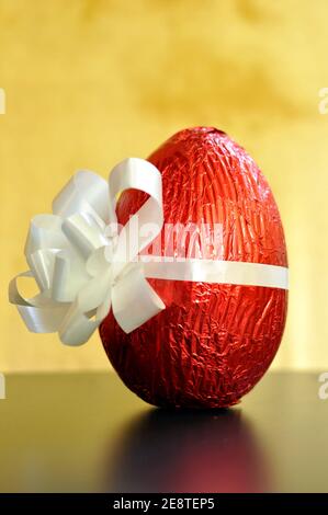 Großes Schokoladenei in rote Folie gewickelt und an einem weißen Knoten für Ostern auf gelbem Hintergrund gebunden. Selektiver Fokus Stockfoto