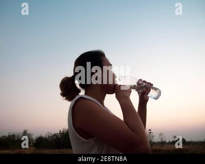 Asiatische Frau Trinkwasser aus Plastikflasche nach dem Training am Abend Sommer auf schönen Sonnenuntergang Himmel Hintergrund. Fitness und gesunde lif Stockfoto