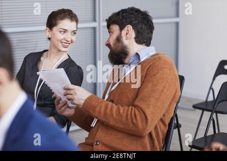 Porträt eines bärtigen Geschäftsmannes, der mit einer lächelnden jungen Frau spricht, während er bei einer Geschäftskonferenz im Publikum sitzt und Raum kopiert Stockfoto