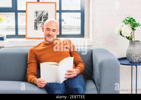 Aufnahme eines gutaussehenden lächelnden Mannes, der ein Buch liest, während er zu Hause auf dem Sofa sitzt. Stockfoto
