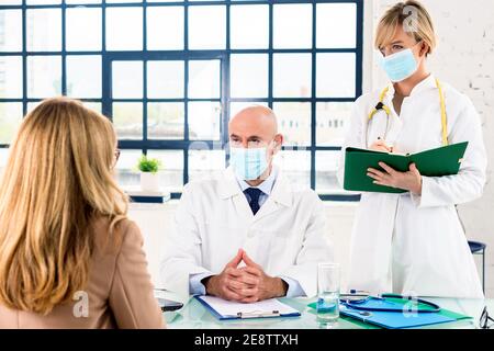 Aufnahme eines männlichen Arztes mittleren Alters, der Gesichtsmaske trägt und mit seinem Patienten spricht, während er am Schreibtisch im Zimmer des Arztes sitzt. Stockfoto