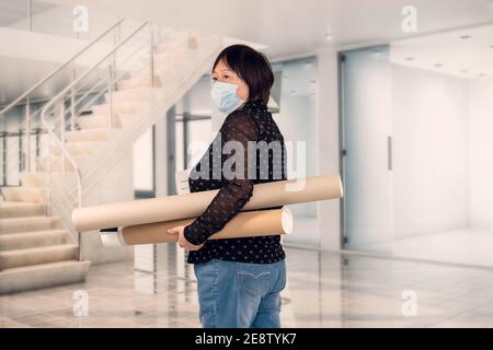 Asiatische Frau mit einer Gesichtsmaske mit einem Ordner und zwei Pappkartonröhrchen in einem Büro. Selektiver Fokus. Arbeits- und Sicherheitskonzept. Stockfoto