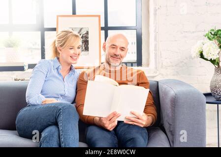 Aufnahme eines glücklichen Paares, das zu Hause auf der Couch sitzt und zusammen Buch liest, während es sich zu Hause entspannt. Stockfoto