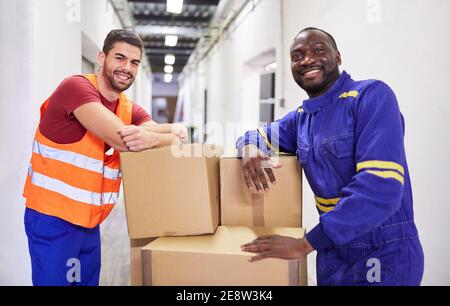 Zwei lächelnde Männer während der Ausbildung als Lagerarbeiter in einem Fabrik mit einem Push-Truck voll von Kartons Stockfoto