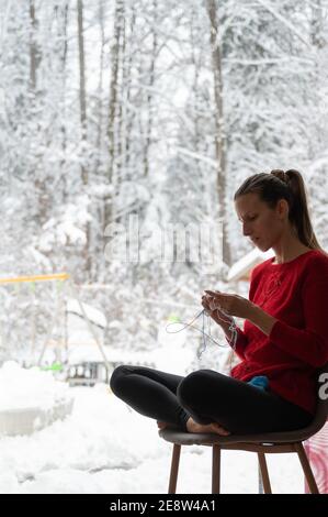 Junge Frau im roten Pullover Stricken mit Baumwollgarn zu Hause, sitzen am Fenster mit verschneiten Natur draußen. Stockfoto