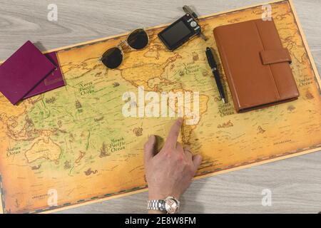 Draufsicht auf die Hand einer Person, die eine Reise vorbereitet und organisiert, die auf einen Punkt auf der Karte zeigt. Pässe, Sonnenbrille, Kamera und Agenda auf Th Stockfoto