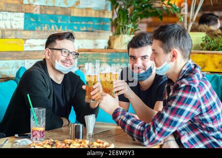 Fröhliche junge Freunde mit Gesichtsmaske trinken Bier im Restaurant Tisch nach dem erneuten Öffnen Stockfoto
