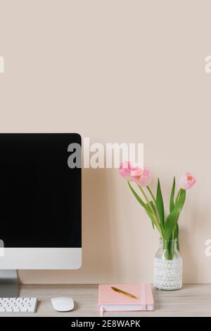 Ein Computermonitor, ein Strauß rosa Tulpen und ein Notizbuch mit einem Stift auf einem hellen Holztisch an einer beigen Wand. Der Arbeitsplatz eines Freiberuflers oder Stockfoto