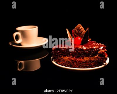 Schokoladenkuchen mit Kirschen und einer Tasse Kaffee auf schwarzem Hintergrund. Kaffee in einer Tasse. Kaffeetasse und Untertasse. Schokoladenkuchen. Lebte mit Kirschen. C Stockfoto