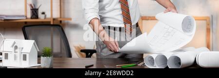 Teilansicht des Architekten Holding Plan in der Nähe Haus Modell und gerollte Blaupausen auf Schreibtisch, Banner Stockfoto