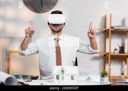 Architect in vr Headset zeigt mit den Fingern, während Sie sitzen auf Arbeitsplatz in der Nähe des Hauses Modell Stockfoto