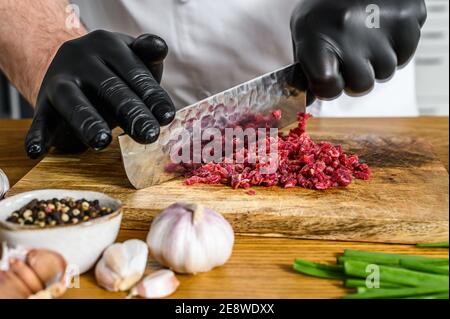 Mann schneidet rohes Rindfleisch Fleisch. Ein Koch in schwarzen Handschuhen. Konzept des Kochens von Rindfleisch Tartare Stockfoto