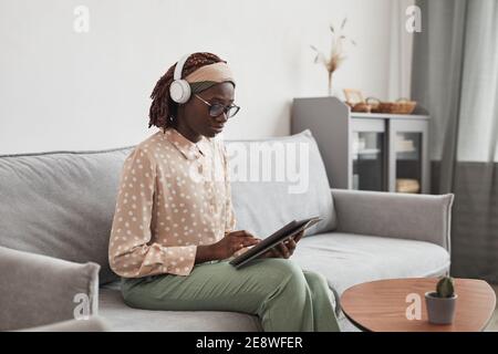 Porträt einer jungen afroamerikanischen Frau, die Kopfhörer trägt und ein digitales Tablet benutzt, während sie zu Hause auf dem Sofa sitzt, im minimalistisch grauen Innenraum, copy-ac Stockfoto