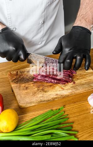 Mann schneidet rohes Rindfleisch Fleisch. Ein Koch in schwarzen Handschuhen. Konzept des Kochens von Rindfleisch Tartare Stockfoto