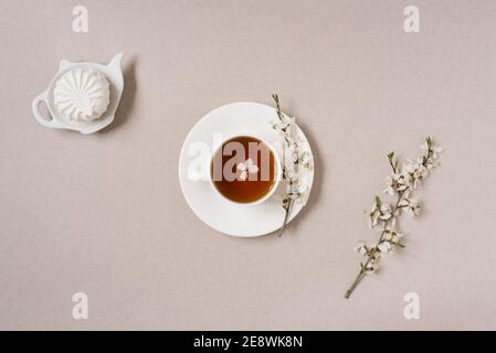 Das Konzept des Frühlings. Eine Tasse schwarzen Tee mit Apfelblüten und Marshmallows auf einer Teekanne Teller auf beigem Hintergrund Stockfoto