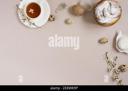 Osterkuchen, Eier, eine Tasse Tee, Süßigkeiten, Zweige eines blühenden Apfelbaums und Federn auf beigem Hintergrund. Platz zum Kopieren, flach liegend Stockfoto