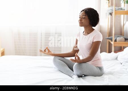 Entspannte schwarze Frau sitzt auf dem Bett und meditiert Stockfoto