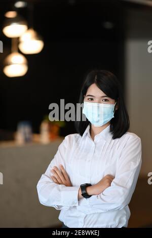 Porträt von selbstbewussten Geschäftsfrau trägt Schutzmaske stehen mit Arm gekreuzt in modernen Büro. Stockfoto