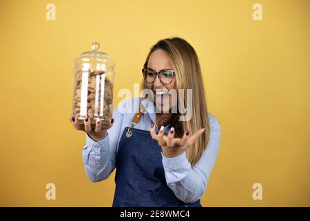 Junge hispanische Frau trägt Bäckeruniform hält ein Cookies-Glas verrückt und verrückt schreien und mit aggressivem Ausdruck und Arme angehoben. Frustr Stockfoto