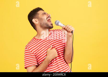 Seitenansicht positive bärtige Mann im gestreiften T-Shirt laut singen Lied hält Mikrofon in der Hand, Spaß in Karaoke ruhen, Sänger Leistung. ICH Stockfoto