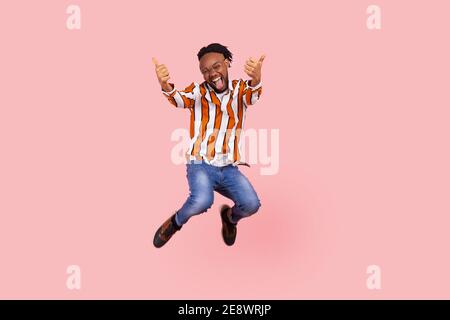 Extrem aufgeregt positiver Afro-amerikanischer Mann mit Dreadlocks im stylischen gestreiften Hemd und Denim-Hose, die Daumen hoch springen lassen, wie fliegen. ICH Stockfoto