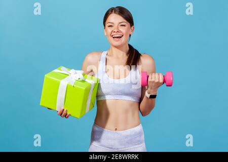 Lächelnd positive Frau in weißer Sportkleidung mit rosa Hantel und große grüne Geschenkbox, erhalten Fitness-Mitgliedschaft im Urlaub. Innenaufnahmen im Studio Stockfoto
