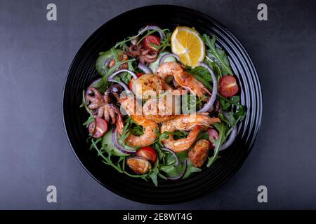 Salat mit Meeresfrüchten, Rucola, Tomaten, Gurken, roter Zwiebel und Zitrone auf einem schwarzen Teller. Schwarzer Hintergrund, Nahaufnahme. Stockfoto