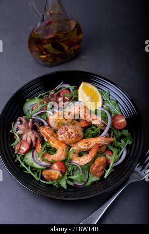 Salat mit Meeresfrüchten, Rucola, Tomaten, Gurken, roter Zwiebel und Zitrone auf einem schwarzen Teller. Schwarzer Hintergrund, Nahaufnahme. Stockfoto