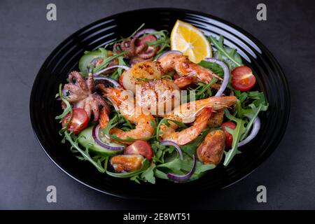 Salat mit Meeresfrüchten, Rucola, Tomaten, Gurken, roter Zwiebel und Zitrone auf einem schwarzen Teller. Schwarzer Hintergrund, Nahaufnahme. Stockfoto