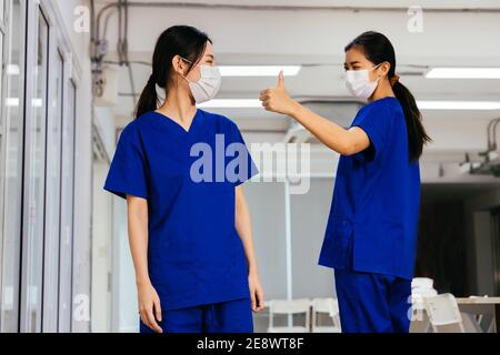 Junge schöne asiatische Krankenschwester in Peeling Uniform trägt Gesichtsmaske bewundern und geben Daumen bis zu Mitarbeiter im Krankenhaus während Covid19 Pandemie. Vertrauen und gute Zusammenarbeit im Konzept der Unternehmenskultur Stockfoto
