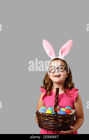Frohe Ostern. Fröhliches kleines Mädchen in einem rosa Kleid mit Tupfen hält einen Korb mit Eiern auf einem grauen Hintergrund. Das niedliche Baby hob seinen Kopf und lo Stockfoto